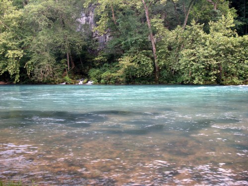 A serene river with turquoise water, surrounded by lush green trees and rocky cliffs in the background.