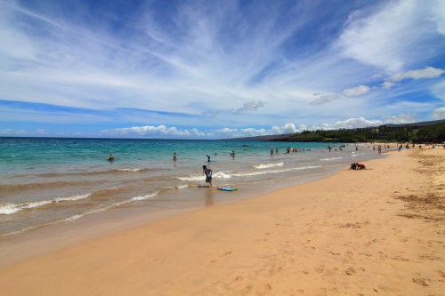 A sunny beach scene with people swimming, surfing, and relaxing on the sand under a blue sky with clouds.