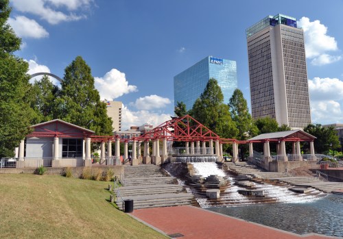 A park with a waterfall feature, surrounded by trees and modern buildings under a blue sky with clouds.