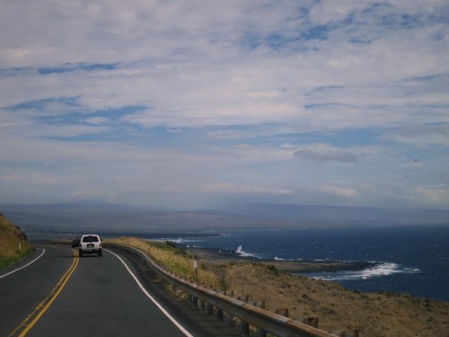 A scenic coastal road curves along the shoreline, with ocean waves and a cloudy sky in the background.