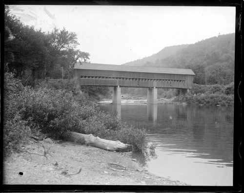 A historic covered bridge spans a river, surrounded by trees and mountains, with a calm water reflection.