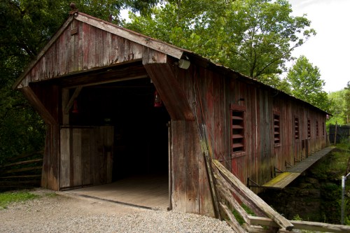 A rustic, weathered covered bridge surrounded by lush greenery and a gravel path leading to its entrance.