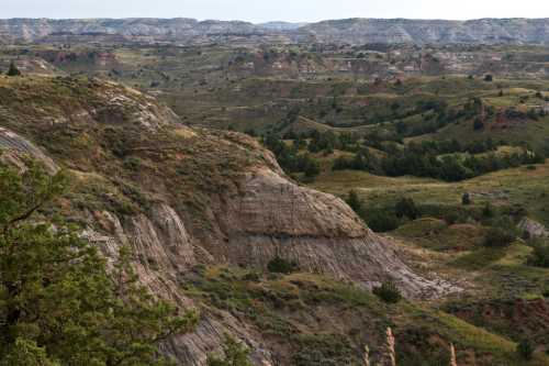 A scenic view of rolling hills and rugged terrain, with green vegetation and layered rock formations under a cloudy sky.