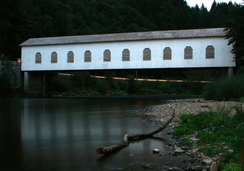 A white covered bridge spans a calm river, surrounded by trees, with a darkening sky in the background.