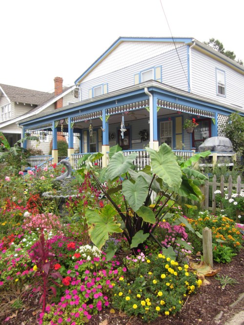 A colorful garden filled with flowers in front of a charming two-story house with a blue porch.