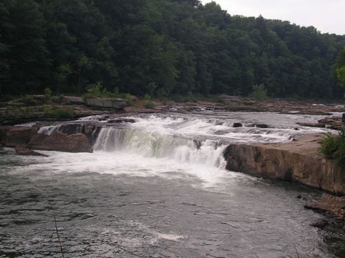 A serene waterfall cascades over rocky ledges, surrounded by lush green trees and a calm river.