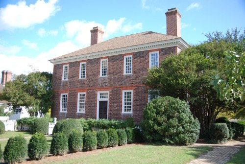 A historic brick house with white windows and a brown door, surrounded by neatly trimmed hedges and a clear blue sky.