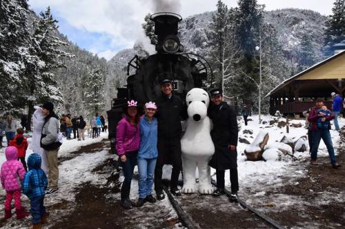 A snowy scene with a steam train, people posing, and a large Snoopy character in front of the train.