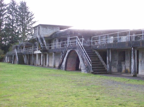 Abandoned concrete building with arched entrances and stairs, surrounded by grass and trees.