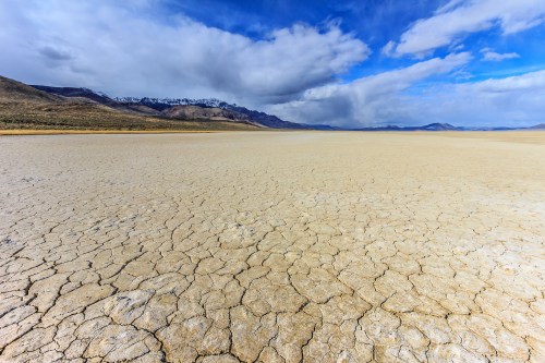 A vast, dry landscape with cracked earth under a blue sky and scattered clouds, mountains in the distance.