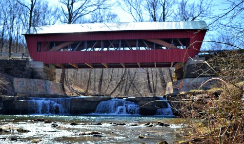 A red covered bridge spans a stream with a small waterfall, surrounded by bare trees and a clear blue sky.