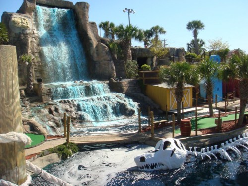 A mini-golf course featuring a waterfall, palm trees, and a dinosaur skeleton in a water feature.
