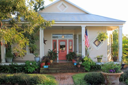 Charming house with a pink door, surrounded by lush plants and an American flag, featuring a welcoming porch.