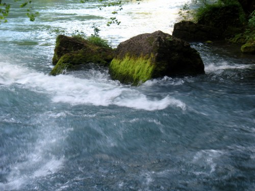 A moss-covered rock partially submerged in a rushing river, with clear blue water flowing around it.
