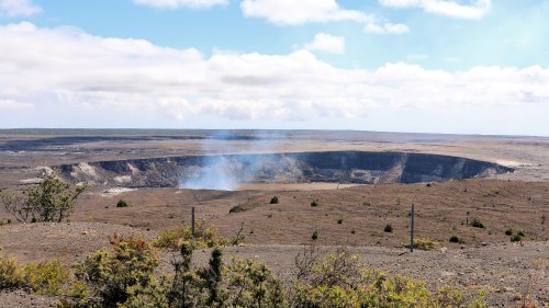 A vast volcanic crater with smoke rising, surrounded by barren land and sparse vegetation under a cloudy sky.