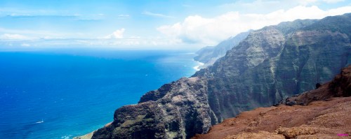 A panoramic view of rugged cliffs meeting a vibrant blue ocean under a clear sky with scattered clouds.
