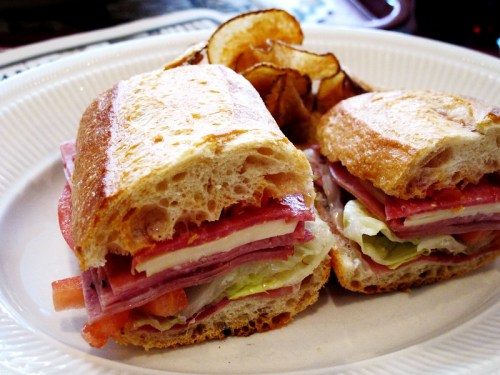 A sandwich with layers of meats, cheese, lettuce, and tomato, served with a side of crispy potato chips.