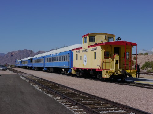 A yellow caboose and blue train cars parked on a railway track with mountains in the background under a clear blue sky.