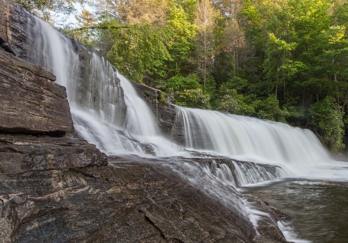A serene waterfall cascades over rocky ledges, surrounded by lush green trees and a tranquil forest setting.