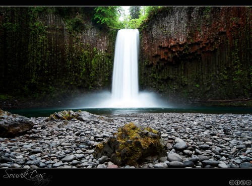 A serene waterfall cascades into a rocky riverbed, surrounded by lush greenery and moss-covered cliffs.