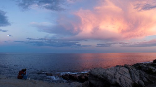 A serene sunset over the ocean, with vibrant pink and purple clouds reflecting on the water, and two people sitting on rocks.