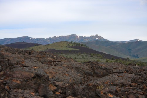 Lava rock foreground with green hills and snow-capped mountains in the background under a cloudy sky.