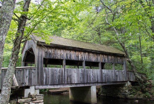A rustic wooden covered bridge spans a small stream, surrounded by lush green trees and foliage.