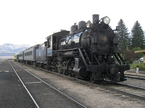 A vintage steam locomotive with a passenger car, parked on tracks with trees and mountains in the background.