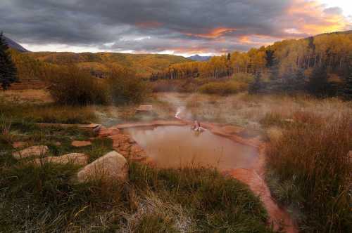 A serene hot spring surrounded by lush grass and trees, with a dramatic sunset sky in the background.