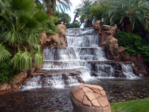A serene waterfall cascades over rocky steps, surrounded by lush greenery and palm trees.