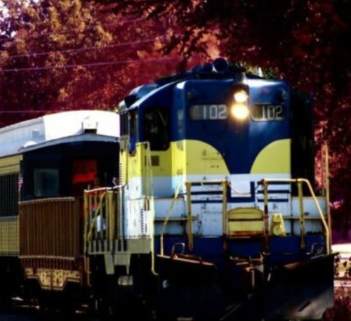 A blue and yellow train engine with the number 102, parked next to a vintage passenger car in a colorful setting.