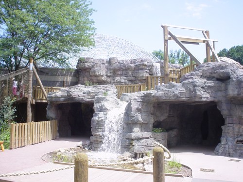 A rocky landscape with a small waterfall, wooden structures, and greenery under a blue sky.