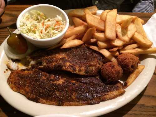 A plate of seasoned fish fillets, golden fries, coleslaw, and two hushpuppies, garnished with a pickle.