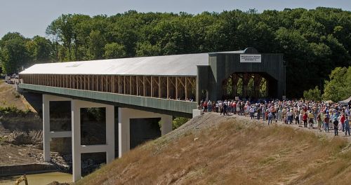 A large covered bridge on a raised structure, with a crowd of people gathered in front, surrounded by trees.