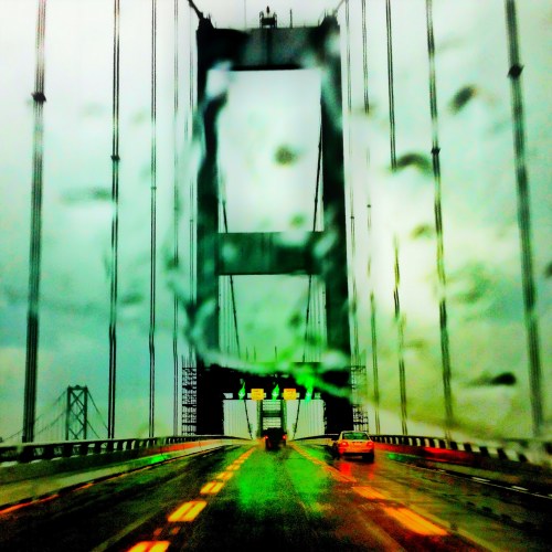 A blurred view of a bridge on a rainy day, with raindrops on the windshield and muted colors.