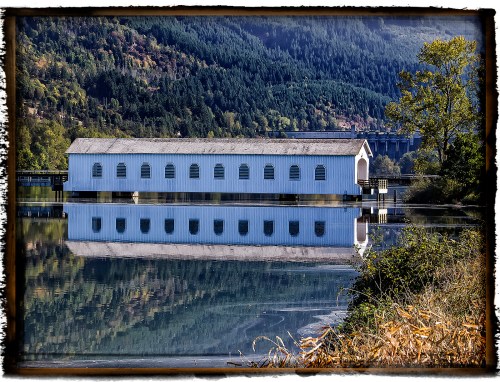 A white building with arched windows reflects in calm water, surrounded by trees and mountains.