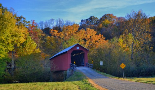 A red covered bridge spans a road, surrounded by vibrant autumn foliage and a clear blue sky.