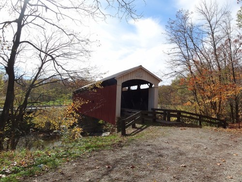 A red covered bridge surrounded by autumn trees and a gravel path under a blue sky with scattered clouds.