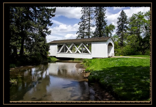 A white covered bridge spans a calm stream, surrounded by lush greenery and tall trees under a blue sky.