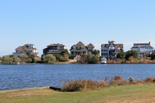 A row of waterfront houses with varied architecture beside a calm lake under a clear blue sky.
