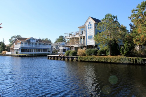 Two waterfront houses with balconies, surrounded by trees, along a calm river under a clear blue sky.
