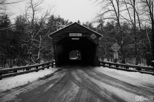 A snow-covered road leads into a rustic wooden covered bridge surrounded by bare trees.