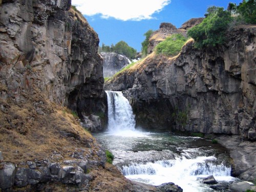 A serene waterfall cascades between rocky cliffs, surrounded by lush greenery and a clear blue sky.