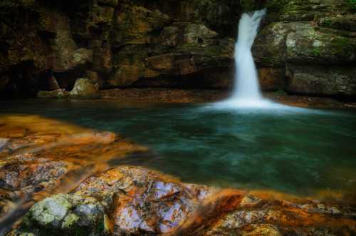 A serene waterfall cascades into a clear pool, surrounded by rocky terrain and lush greenery.