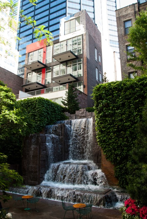 A modern building with balconies overlooks a cascading waterfall surrounded by greenery and urban architecture.