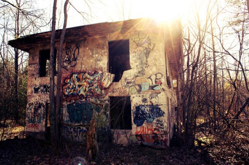 Abandoned building covered in graffiti, surrounded by trees, with sunlight shining through the top.