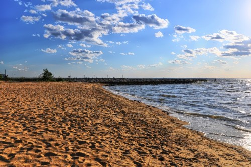 A sandy beach with gentle waves, under a blue sky dotted with clouds, and a distant rocky jetty.