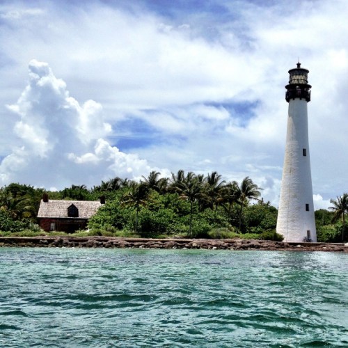A tall white lighthouse stands beside a small building, surrounded by palm trees and blue skies with fluffy clouds.