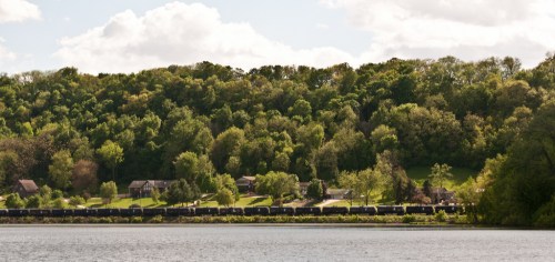 A serene landscape featuring a lake, lush green hills, and a train passing through a tree-lined area with houses.