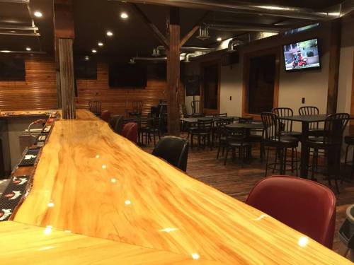 A wooden bar with a polished surface, surrounded by black chairs and tables, in a cozy, dimly lit interior.
